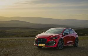 Vista frontal tres cuartos del nuevo Renault Clio VI de color rojo aparcado en un paisaje rural al atardecer.