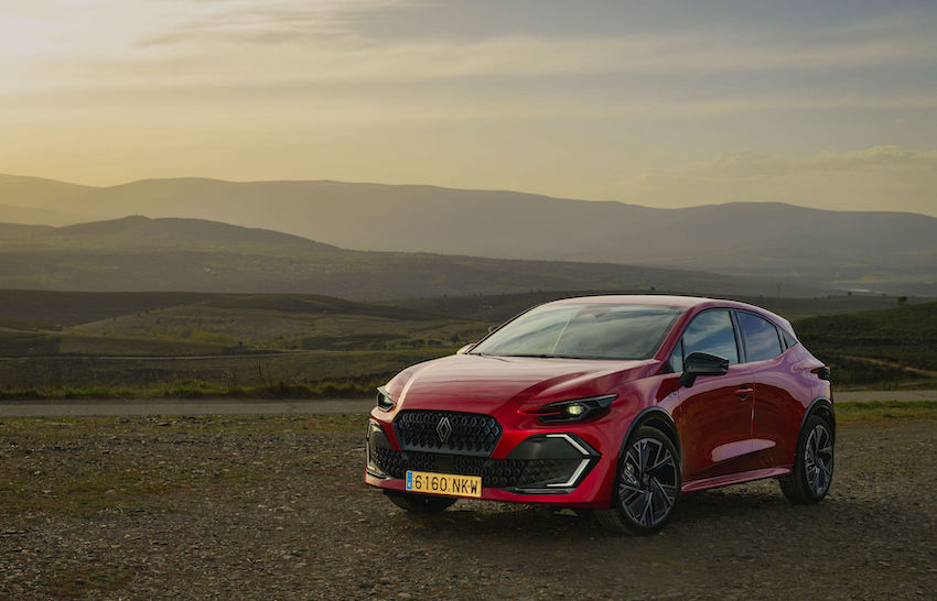 Vista frontal tres cuartos del nuevo Renault Clio VI de color rojo aparcado en un paisaje rural al atardecer.